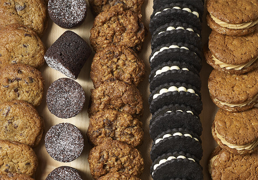 A wooden platter with several rows of cookies and a chocolate brownie on it.