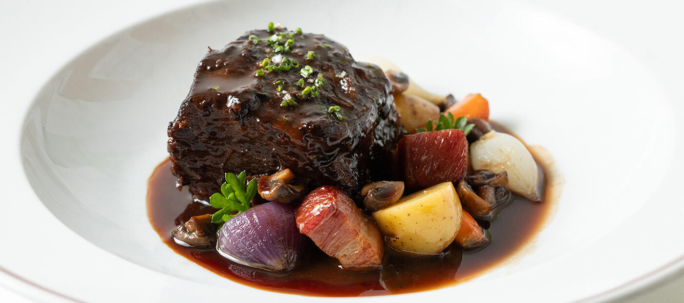 A shallow dish containing boeuf bourguignon, with bacon lardons, onions, potatoes, carrots and topped with chives.